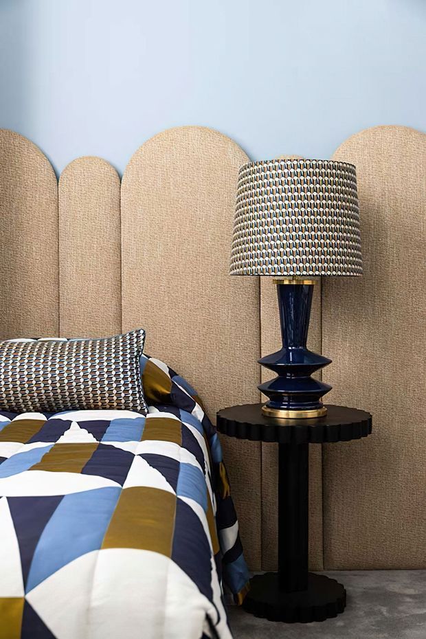 A stylish bedroom corner featuring a bed with bold geometric blue, white, mustard, and black bedding and a patterned matching cushion. A sculptural dark navy table lamp with a patterned drum shade sits on a black fluted pedestal side table. Behind the bed is a curved, upholstered tan headboard or room divider, set against a pale blue wall and grey carpet.