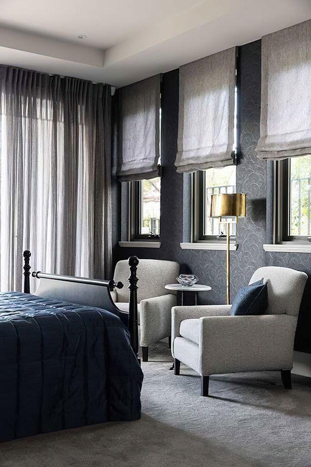 A sophisticated bedroom with a dark patterned feature wall, three windows dressed in textured Roman blinds, and full-height sheer curtains. A black metal bed with deep navy bedding sits beside two light upholstered armchairs, a small round side table, and a brass floor lamp. The room has soft grey carpeting, layered window treatments, and a refined, moody atmosphere.