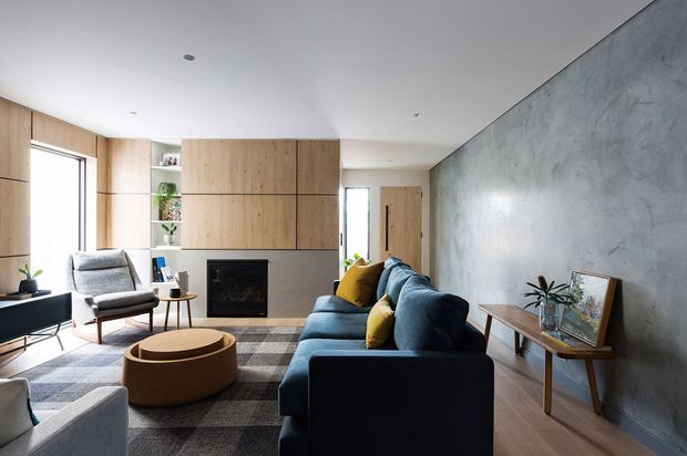 Contemporary living room with a dark sectional sofa, patterned rug, rounded upholstered coffee table, and mid-century-style lounge chair. Full-height timber cabinetry incorporates an open shelf and black fireplace, while a textured grey concrete-look feature wall contrasts with the warm timber floor and joinery. The room is bright, restrained, and refined with layered neutral furnishings and small yellow and green accents.