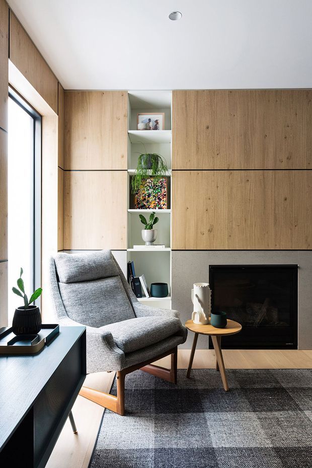 A compact, light-filled living room with a grey upholstered lounge chair, small round timber side table, black media console, and a dark fireplace set into a textured grey wall. Full-height light timber cabinetry frames open shelving with books, artwork, and plants. A large grey checked rug and pale timber flooring create a calm contemporary interior.