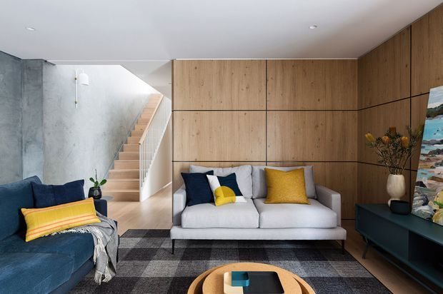 Contemporary living room with a grey sofa and navy armchair arranged around a low coffee table, set against warm timber wall panelling. A concrete-textured wall and open staircase add architectural interest, while mustard and blue cushions provide colour accents. The room has recessed ceiling lighting, a dark rug, built-in dark cabinetry, and artwork.