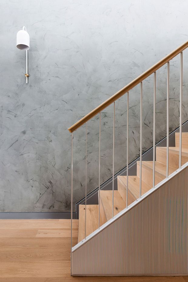 A contemporary stairway with pale timber stairs and flooring, slim vertical metal balusters, and a warm timber handrail. The stair side is clad in vertically fluted timber, while the surrounding wall has a textured grey plaster finish. A simple white wall light provides a small accent.
