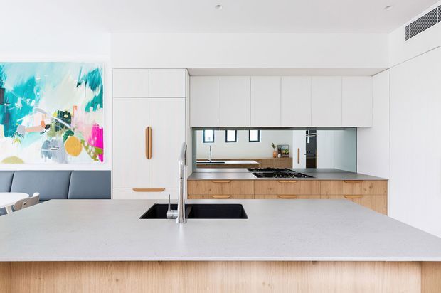 Bright open-plan kitchen viewed from the island, with a large pale stone island benchtop and integrated sink, white full-height cabinetry, light timber lower cabinetry, a mirrored splashback, and a compact gas cooktop. A dark blue built-in banquette and colourful abstract artwork are visible to the left. The space has a clean, contemporary fit-out with recessed lighting and concealed air conditioning.