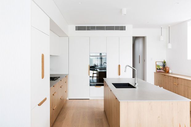 Bright, refined contemporary kitchen with a large central island, pale timber cabinetry, white full-height joinery, integrated appliances, a dark undermount sink, and brushed metal tapware. The room has a light timber floor, white walls and ceiling, streamlined pendant lighting, and a warm minimalist palette.