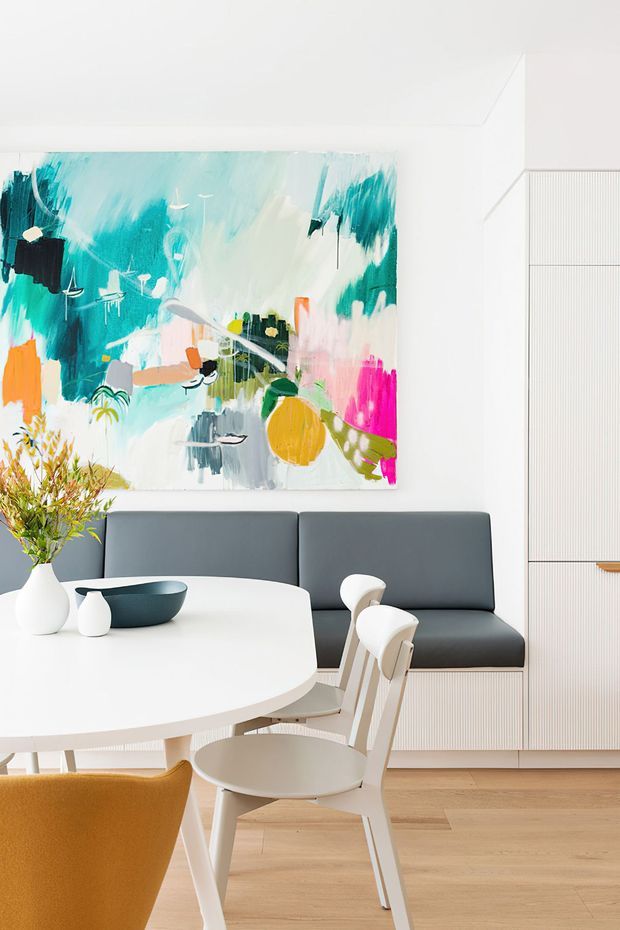 Bright contemporary dining area with a large white oval table, sculptural light wood and white dining chairs, dark grey built-in banquette seating, pale timber flooring, and ribbed white cabinetry or wall panelling. A large colourful abstract artwork provides a vivid turquoise, pink, orange and green focal point, with simple ceramic vessels and foliage on the table.