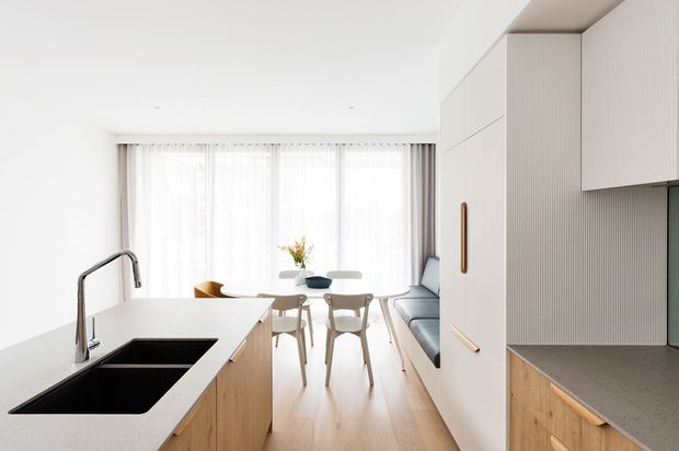 Bright, open-plan contemporary kitchen and dining area with a large white island, black double sink and brushed metal tap, pale timber cabinetry and flooring, and a tall integrated storage wall. A compact dining table sits beside full-height sheer-curtained glazing, with a grey sofa visible beyond the kitchen.