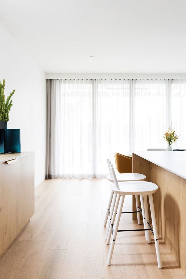 A bright, minimalist kitchen or dining area with white walls and ceiling, pale timber flooring, and a long timber kitchen island or breakfast bar. Two white and timber stools sit at the island, while full-height sheer curtains provide abundant filtered daylight. A pale timber sideboard with blue planters is visible on the left, and a small vase of foliage decorates the benchtop.