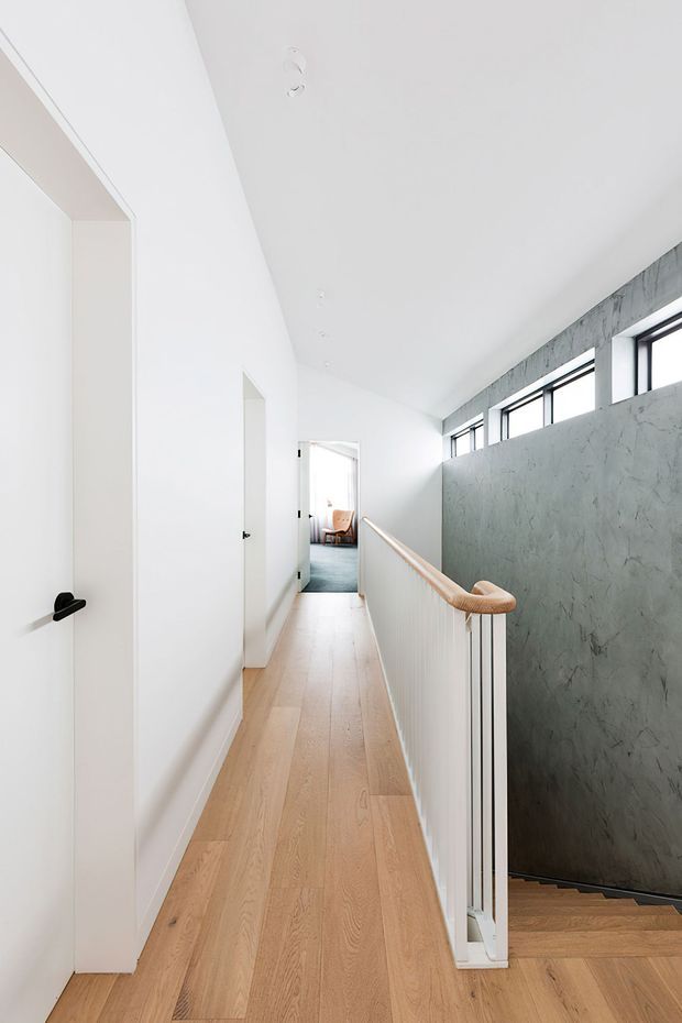 Bright minimalist upper-level hallway with pale timber flooring, white walls and ceiling, a stair opening with a slim timber handrail and white balustrade, and a textured grey feature wall beside high horizontal windows. A bedroom or sitting area is visible at the far end.