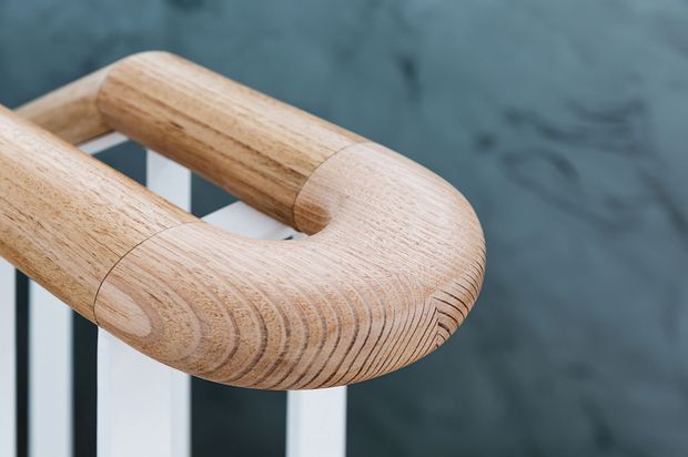 Close-up detail of a rounded, laminated timber handrail curving around a stair or landing corner. The light natural wood shows prominent grain and layered joinery, with white vertical balusters and a dark grey wall in the background.