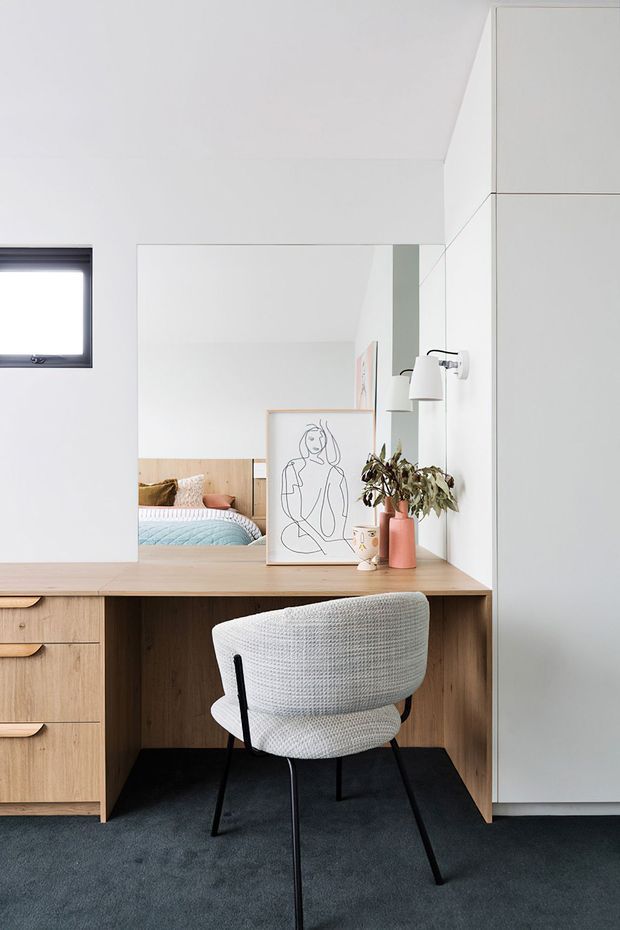 A compact, minimalist bedroom workspace with a built-in timber desk and drawers, wall-to-wall mirror, upholstered chair, wall-mounted lamp, framed line artwork, and a small vase of foliage. The mirror reflects a bed with pale blue bedding and timber wall panelling, confirming the workspace is within a bedroom.