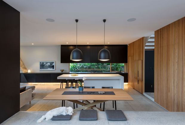 Open-plan contemporary kitchen, dining, and living space with dark integrated cabinetry, a large pale island, timber dining table, and two oversized black pendant lights. Vertical timber slats form a feature wall and screen along the right side, while a narrow glazed opening behind the kitchen provides greenery views. The palette is restrained and warm, combining charcoal, pale timber, concrete-like flooring, and soft neutral furnishings.