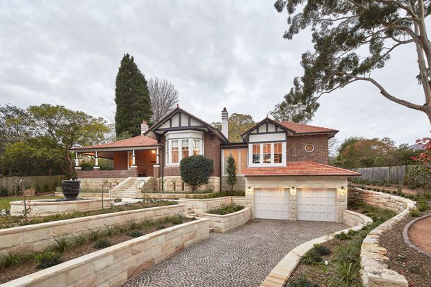 Exterior view of a substantial brick residence with gabled roof forms, red terracotta roof tiles, white-trimmed projecting bays, and a covered verandah. The property features terraced sandstone retaining walls, landscaped garden beds, a curved cobblestone driveway, and an integrated double garage. The traditional detailing and mature landscaping create a refined suburban character under an overcast sky.