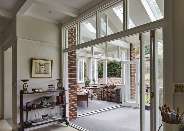 A bright transitional interior looking through a large glazed opening into an enclosed sunroom or conservatory. The space features extensive white-framed glazing, brick structural piers, a grey carpeted floor, pale walls, a coffered ceiling, and traditional dark timber display shelving with decorative objects. The adjoining glazed room contains casual seating and a small table, with a garden visible beyond.