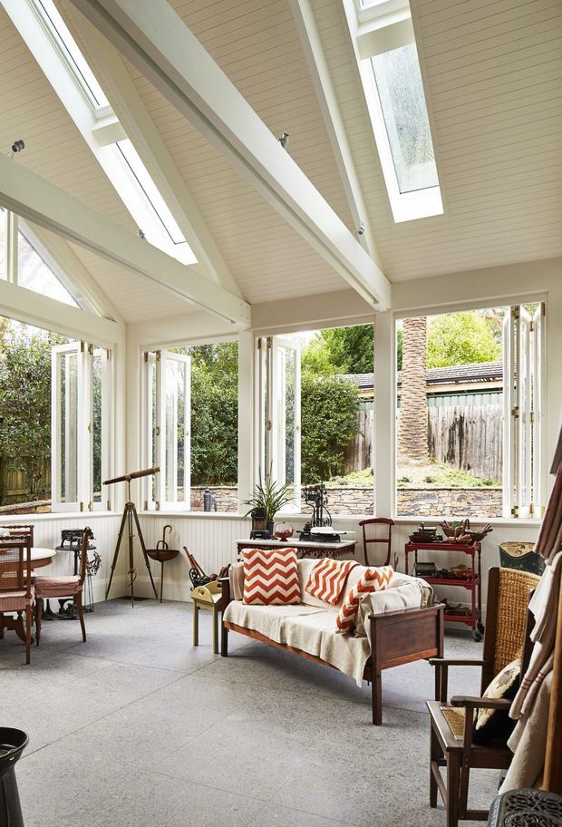 Bright, airy sunroom or conservatory with a high pitched, white-painted timber-lined ceiling, exposed beams, multiple skylights and extensive bi-fold or casement glazing. The room is furnished as a relaxed sitting area with a vintage wooden sofa, patterned cushions, armchairs, a small table, telescope and decorative furniture. Pale grey flooring, white wall panelling and abundant garden views create an inviting indoor-outdoor atmosphere.