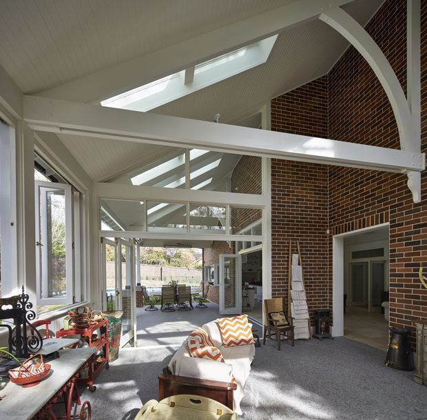 A bright, spacious glazed indoor-outdoor living area with a high vaulted white-lined ceiling, multiple skylights and clerestory glazing, exposed white structural beams, and a prominent dark red-brown brick feature wall. The room contains casual seating, a small bar cart, a fireplace area, and outdoor views through extensive windows and doors.