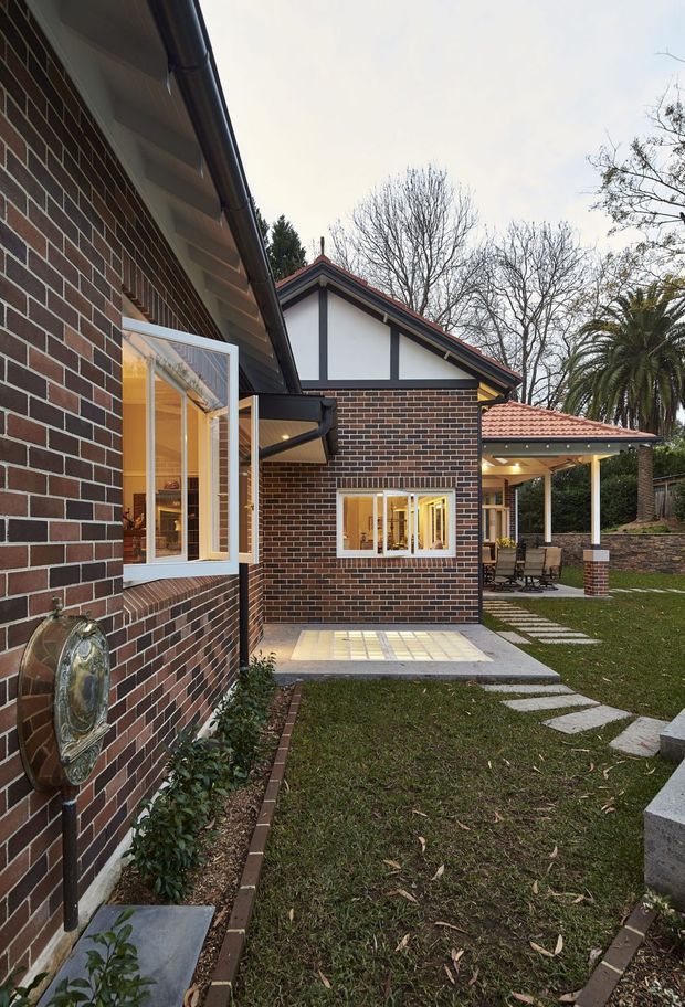 Exterior view of a traditional brick residential property, showing a landscaped courtyard or side garden between connected brick volumes. The scene includes dark red-brown brick walls with contrasting white gable infill and dark timber-like trim, white-framed opening windows, tiled pitched roofs, gutters and downpipes, and a covered outdoor entertaining area with a terracotta-tiled roof. Stepping-stone paths cross the lawn, with low brick garden edging and a stone retaining wall. The image is photographed at ground level in an overcast setting.