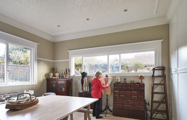 A bright, traditionally detailed room functioning as a dining and display space. It features a large light timber dining table, extensive windows, taupe walls with white trim and dado moulding, ornate pressed-pattern ceiling detailing, decorative cornices, a dark timber vintage apothecary-style chest, display objects, a mannequin, and a leaning timber ladder. The room has abundant natural light and a curated, eclectic vintage atmosphere.