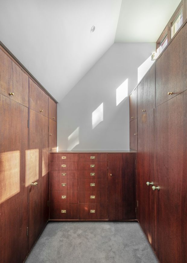 Compact walk-in wardrobe lined with dark reddish-brown timber cabinetry on both sides and across the rear wall. The fitted storage includes tall cupboard doors, numerous drawers with small brass pulls, and a grey carpeted floor. A high clerestory window admits strong sunlight onto the white walls, creating a bright, warm contrast with the dark joinery.