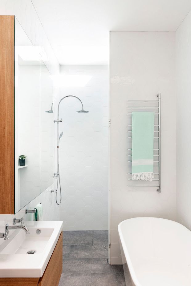 Bright, compact contemporary bathroom with a walk-in shower, freestanding white bathtub, wall-mounted heated towel rail, mirrored cabinetry, and a timber vanity. White hexagonal shower wall tiles contrast with grey floor tiles, while chrome fixtures and pale aqua towels add subtle detail.