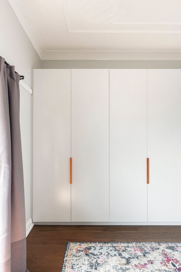 Bright bedroom corner featuring a full-height wall of white built-in wardrobes with slim warm timber handles, dark timber flooring, a patterned area rug, mauve curtains, and ornate decorative ceiling cornices and ceiling detailing.
