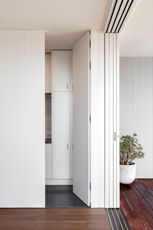A bright, minimalist interior transition space with extensive white vertical-panel cabinetry and folding/sliding doors. The view looks toward a compact storage or utility area with tall built-in cupboards, dark flooring, and a glimpse of a kitchen-like work surface. At right, open glazed doors connect to a timber deck, with a potted plant adding natural contrast. Warm timber flooring runs through the foreground, while slim black door tracks and hardware provide linear accents.