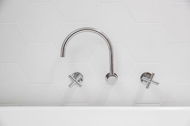 Close-up of a minimalist bathroom wall featuring a chrome curved-spout tap with separate cross-handle controls, set against white hexagonal wall tiles. The white edge of a bathtub or basin is visible along the bottom.