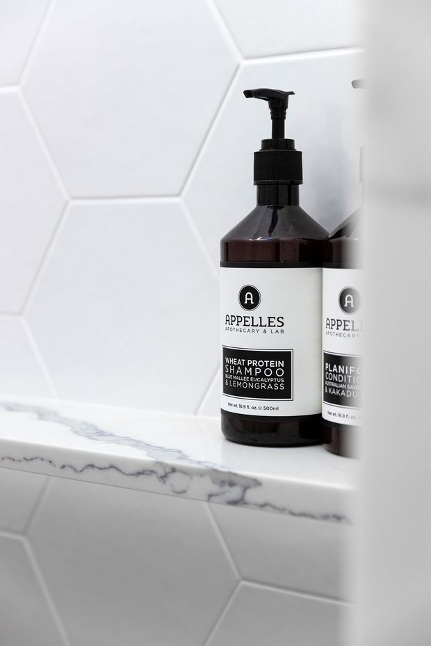 Close-up product-style photograph of two dark amber pump bottles of Appelles shampoo and conditioner placed on a white marble-edged tiled bathroom shelf. The background features large white hexagonal wall tiles and matching white geometric tiling below, with the bottles reflected in a mirror on the right.