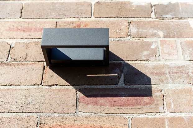 Close-up exterior detail of a dark rectangular wall-mounted light fixture attached to a weathered pale brick wall. Strong sunlight casts a defined shadow across the brickwork, highlighting the fixture's geometric form.