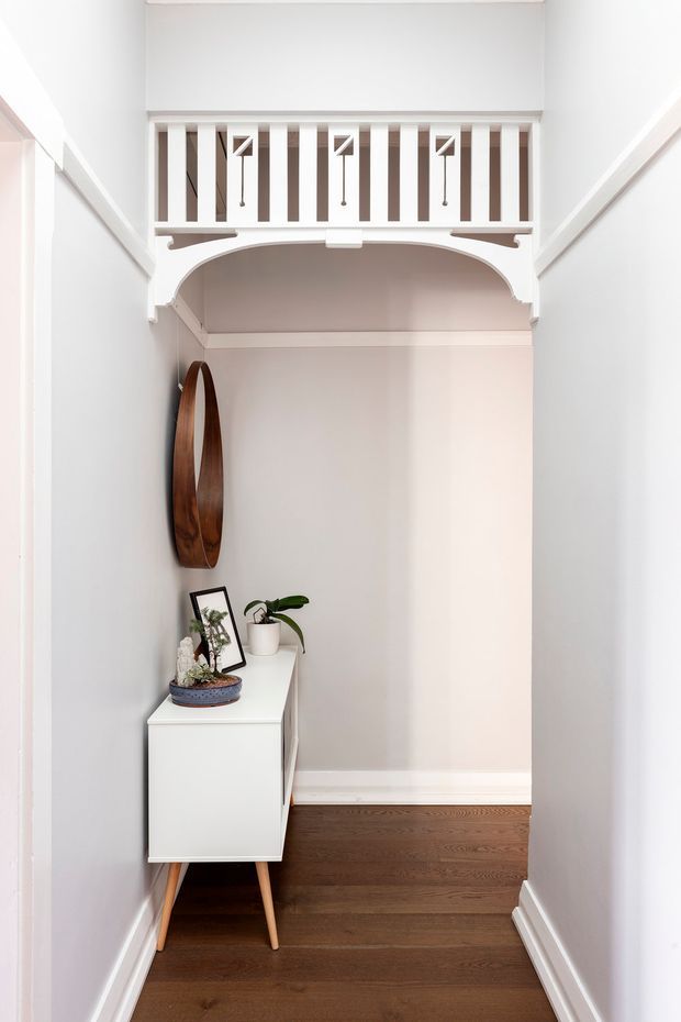 A narrow, bright residential hallway or entry passage with white walls and trim, dark timber flooring, and a small white mid-century-style console on tapered wooden legs. A round wooden wall mirror and potted plants decorate the console. At the end of the passage is an ornate white arched opening with a balustrade or mezzanine railing above, featuring vertical spindles and small hanging light fixtures.
