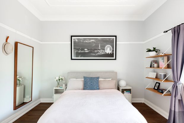 A bright, neatly styled bedroom with a centered upholstered bed, pale pink bedding, and a blue patterned accent cushion. The room has light grey walls with white trim and decorative ceiling moulding, dark timber flooring, a large timber-framed wall mirror, open timber shelving, a black-and-white framed artwork, and purple curtains.