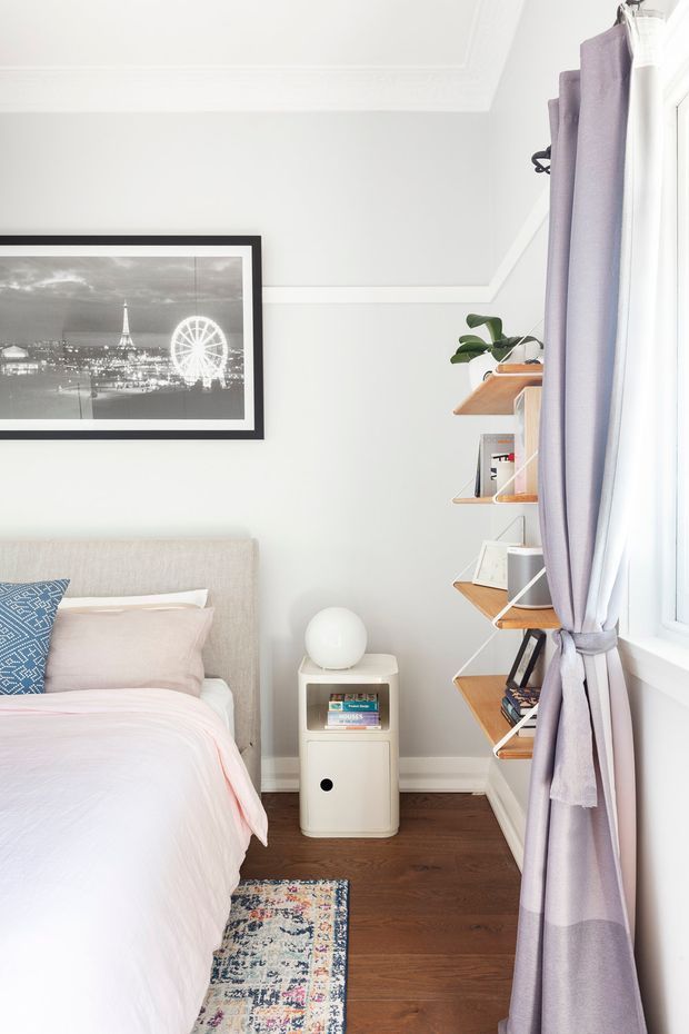 Bright, compact bedroom corner with a partially visible upholstered bed dressed in pale blush and white bedding, a blue patterned accent pillow, dark timber flooring, and a small patterned rug. A white bedside cabinet with a spherical lamp sits beside the bed, while open timber wall shelving provides storage and display. Full-height windows with tied-back lavender curtains bring in abundant daylight. The room has a clean, softly coloured, contemporary residential interior.