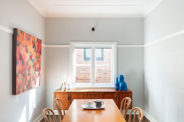 Bright, compact dining room with a rectangular timber dining table and bentwood chairs, a warm timber sideboard beneath a central window, and a colourful abstract artwork on the left wall. The room has pale grey walls, white decorative cornices and dado-style trim, a patterned ceiling detail, and strong natural daylight. A brick building is visible through the window.
