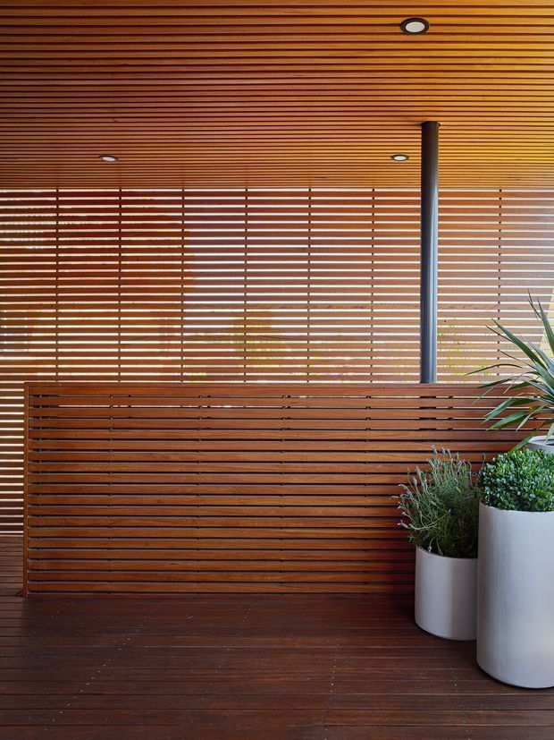 A warm, timber-lined covered outdoor space with horizontal slatted screening across the rear and ceiling, a dark timber deck floor, recessed downlights, and a slim dark structural post. Large white cylindrical planters with green foliage occupy the right side. The space has a refined, contemporary indoor-outdoor character.