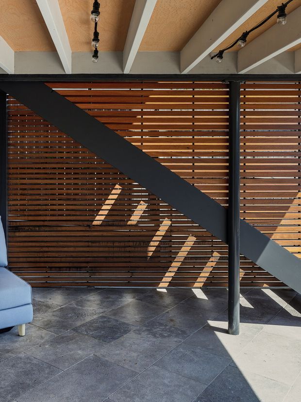 A partially enclosed contemporary indoor-outdoor space with dark grey tiled flooring, a warm timber horizontal slatted privacy screen, exposed structural beams and black steel supports. A prominent diagonal black brace crosses the view, with exposed track lighting on the timber-toned ceiling. Sunlight casts strong linear shadows across the floor and wall; the edge of a pale upholstered chair is visible at left.