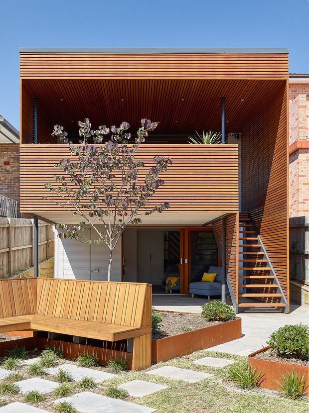 Exterior view of a compact multi-storey contemporary residence arranged around a landscaped courtyard. The facade, soffit, balcony privacy screen and stair enclosure are clad in closely spaced horizontal timber battens, with a deep upper-level opening and exposed external stair. A built-in timber bench, concrete stepping slabs, raised rust-coloured planter beds and drought-tolerant planting occupy the foreground. Brickwork from adjacent or adjoining walls is visible at the sides.