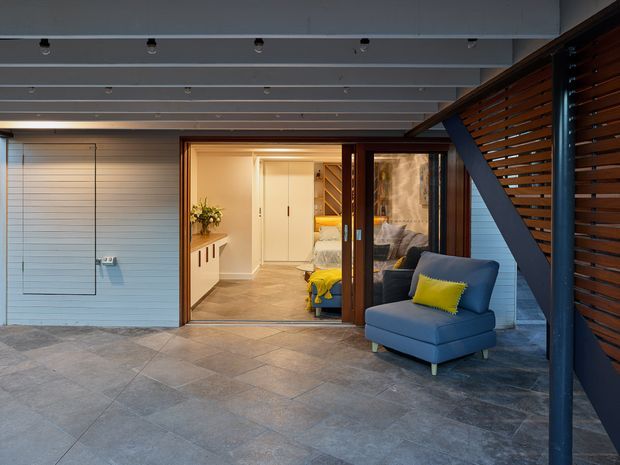 Covered outdoor terrace or courtyard looking toward an open glazed entry into a compact interior. The space features grey tiled paving, white horizontal wall cladding, exposed painted structural beams with recessed spotlights, dark timber slatted screening, and a blue upholstered armchair with a bright yellow cushion. Through the open doors, a small living or bedroom area and white built-in cabinetry are visible.