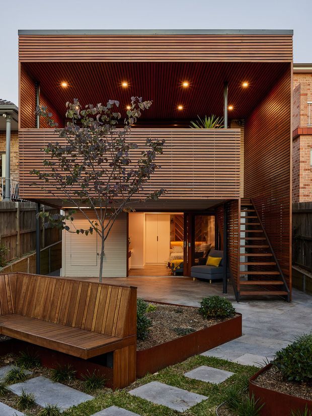 A compact two-storey residential extension viewed from a landscaped courtyard. The facade and upper-level soffit are clad in closely spaced horizontal timber battens, with a covered outdoor area, integrated planter beds, a built-in timber bench, and an open external stair leading to the upper level. Warm recessed lighting illuminates the timber-lined terrace and glazed living area.