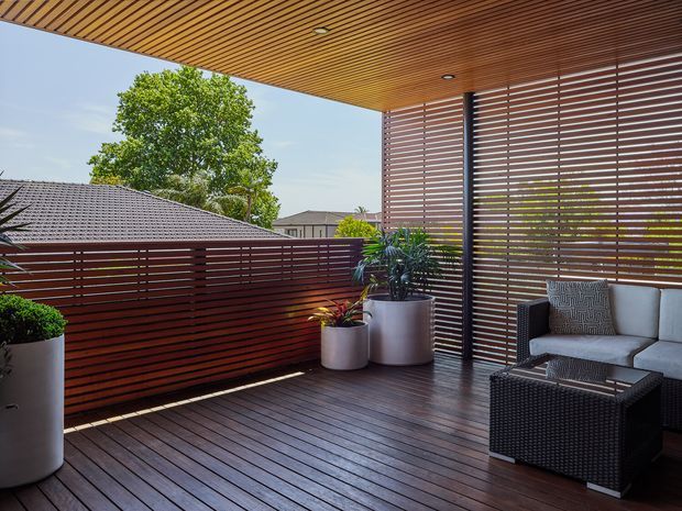 A covered outdoor terrace with dark timber decking, a slatted timber ceiling, and horizontal timber privacy screens forming the balustrade and side wall. Potted plants and a contemporary outdoor lounge setting with a grey sofa, patterned cushions, and black woven coffee table occupy the space. The terrace overlooks neighbouring rooftops and mature trees in bright daylight.