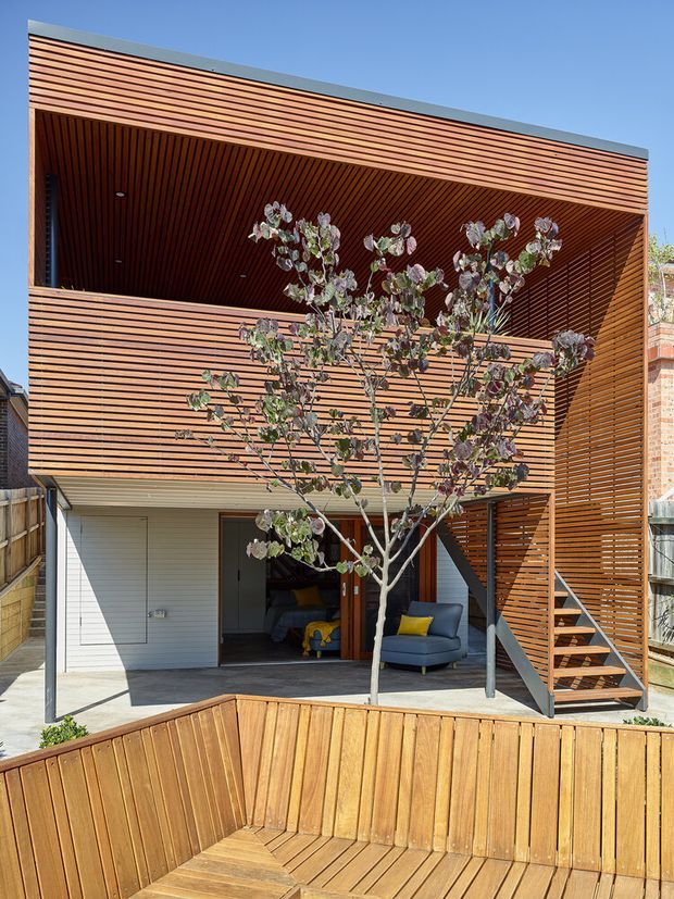 Exterior view of a contemporary multi-storey home with extensive horizontal timber slat cladding and a deep recessed upper balcony lined in timber. The ground level opens to a small outdoor seating area, while an external timber stair leads to the balcony. A timber bench and planter form the foreground, with a sculptural tree partially screening the facade.