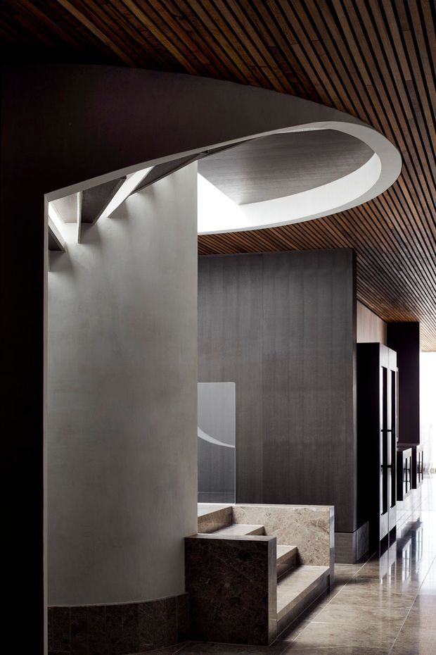 Dramatic contemporary interior corridor or stair hall featuring a sculptural curved white ceiling opening, dark grey wall panels, a richly slatted timber ceiling, stone-clad stair and low walls, and polished stone flooring. Strong linear perspective and daylight at the far end emphasize the architectural composition.
