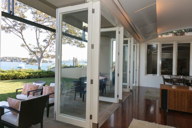 A bright enclosed veranda or sunroom with full-height bi-fold glazed doors opening onto a lawn and waterfront garden. The room has polished dark timber flooring, a white-lined ceiling, white-framed windows, and a compact dining area, while outdoor lounge seating is visible beyond the open doors.
