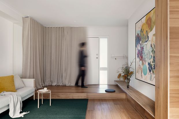A bright, compact contemporary living room and entry area with a raised timber platform, built-in timber cabinetry and shelving, white walls, and full-height neutral curtains. A white sofa with a mustard cushion sits beside a small round side table, while a large colourful artwork and simple floral arrangement add visual interest. A frosted-glass entry door and blurred person are visible in the background.