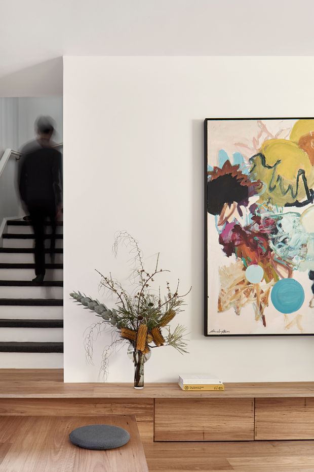 A contemporary minimalist living area with a warm timber built-in media console and matching coffee table. A large colourful abstract artwork is mounted on the white wall, while a sculptural dried floral arrangement and books decorate the console. An open stairway with dark treads is visible at left, and a blurred person is ascending it.