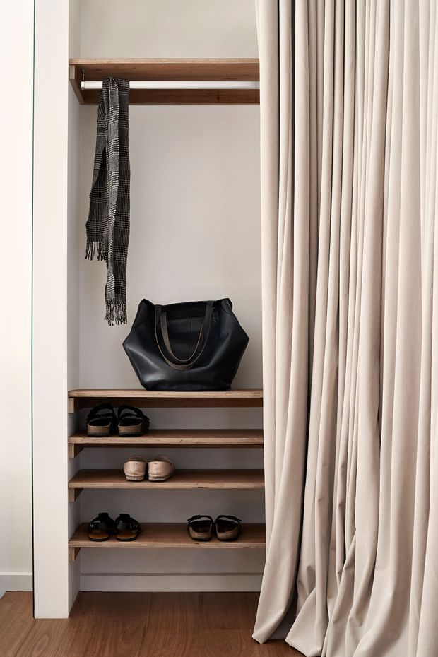 A built-in open wardrobe or dressing alcove with warm timber shelves, a hanging rail, folded or hanging accessories, shoes, and a black handbag. Full-height soft cream curtains partially screen the storage, while dark timber flooring and neutral walls create a refined, understated interior.