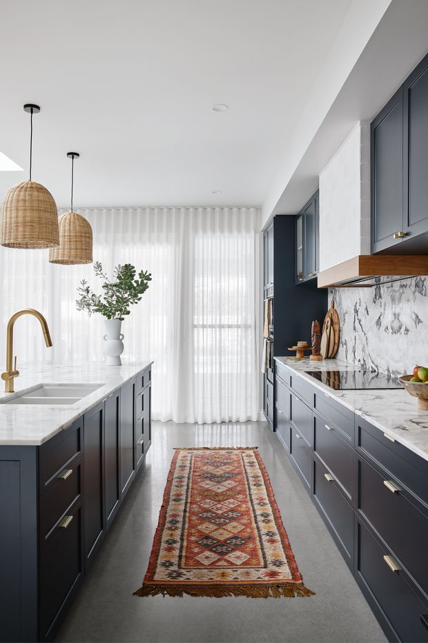 A bright, narrow galley-style kitchen with dark navy shaker cabinetry on both sides, white marble-look benchtops and splashback, a central island with an undermount sink and brushed brass tapware, woven pendant lights, sheer full-height curtains, and a patterned runner rug. The space combines refined cabinetry with warm natural accents and abundant diffused daylight.