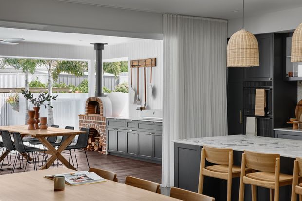 Open-plan kitchen and dining area with a large marble-look island, dark grey cabinetry, timber dining furniture, woven pendant lights, and a white vertical-panelled wall. A built-in brick pizza oven and outdoor garden are visible beyond the dining zone, creating a bright, contemporary family entertaining space.