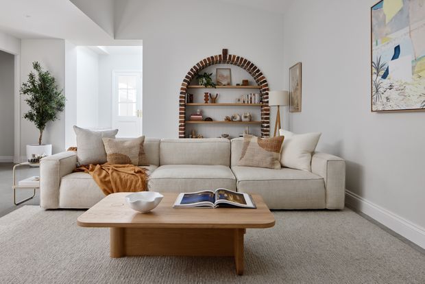 Bright, refined living room with a large neutral sectional sofa, textured cushions, a warm timber coffee table, and a built-in arched brick-lined shelving niche displaying books and decorative objects. White walls, pale carpet, soft natural light, indoor greenery, and abstract artwork create a calm contemporary setting.