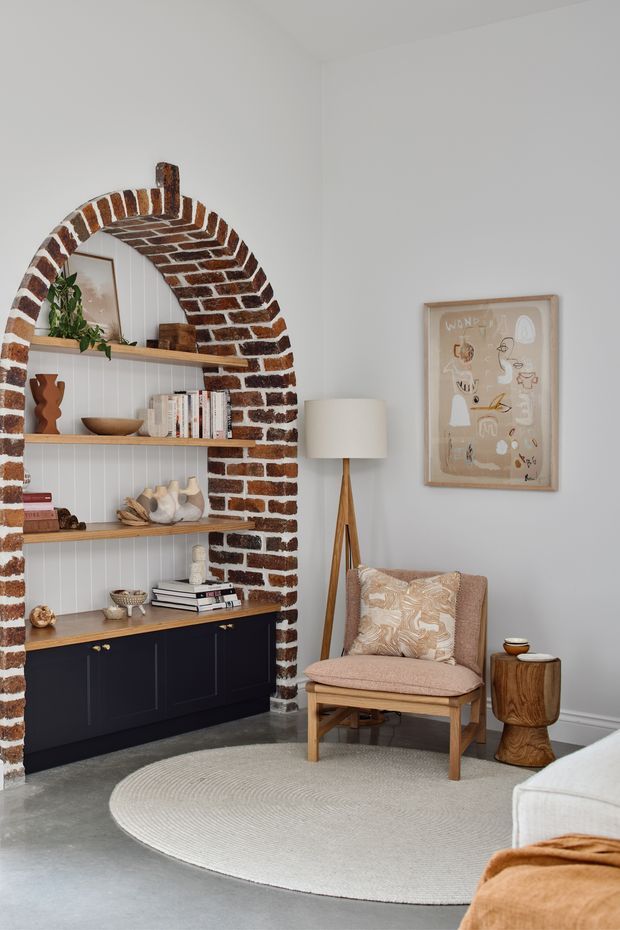 A cozy living room corner featuring a built-in arched brick shelving alcove with timber shelves and dark cabinetry below. The space includes a wood-framed upholstered armchair, tripod floor lamp, sculptural timber side table, framed artwork, and a round textured rug. The palette is soft and neutral with warm wood and exposed brick accents.
