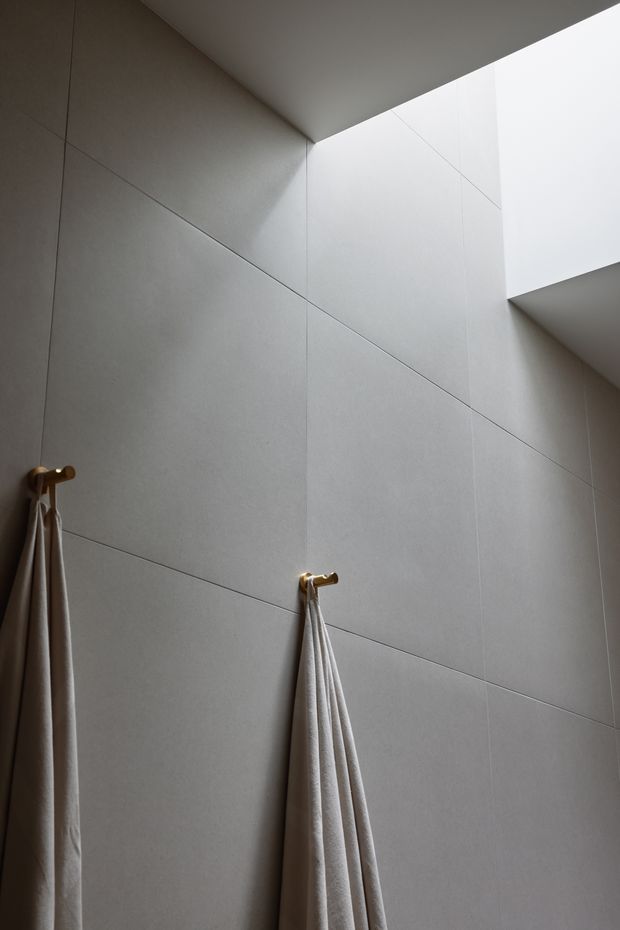 Minimalist bathroom interior featuring large-format light grey wall tiles with fine grout lines, two brass towel hooks with hanging neutral towels, and a high-level skylight or clerestory opening that brings in soft natural light. The composition emphasizes clean geometry, subdued tones, and vertical scale.