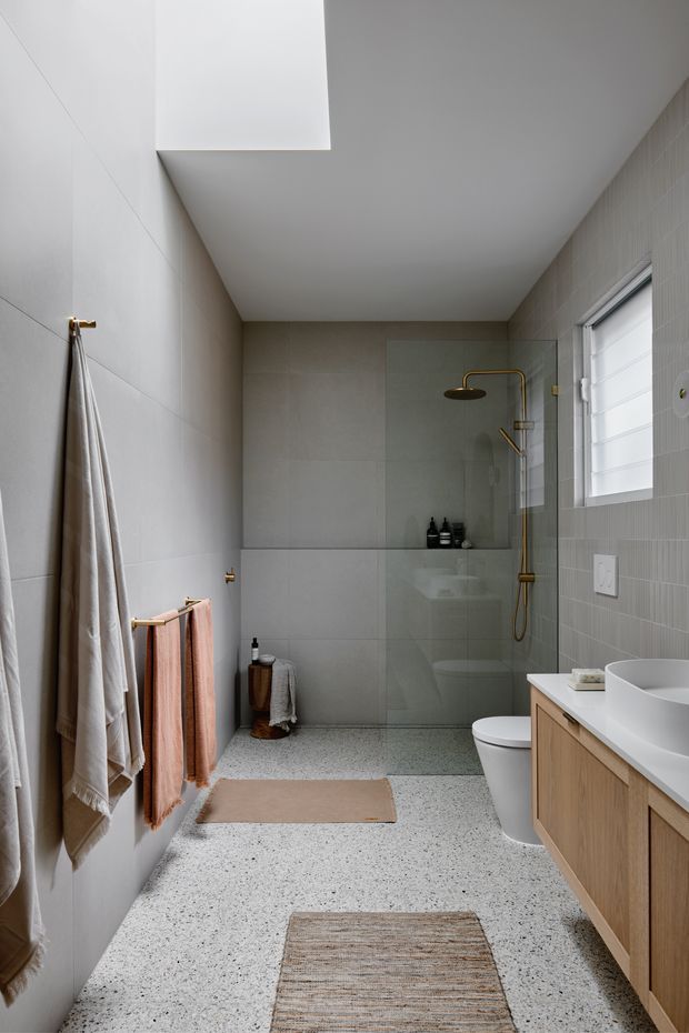 A narrow contemporary bathroom with a walk-in shower enclosed by a frameless glass screen, wall-mounted rain shower and brushed brass fixtures. The room features pale large-format tiled walls, a terrazzo-style floor, a timber vanity with a white vessel basin, a white toilet, and soft neutral towels. A high horizontal window provides natural light.